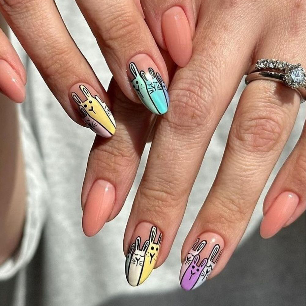 Almond-shaped peach nails featuring detailed cartoon bunny nail art design. Colorful pastel rabbits in yellow, mint, and purple are painted on accent nails. Simple peach polish on others, with a diamond ring.